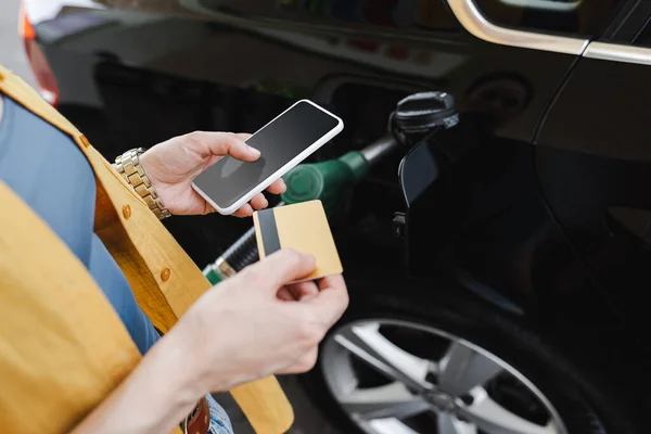 ガソリンスタンドで車を給油しながらスマートフォンやクレジットカードを持っている女性の作物ビュー — ストック写真