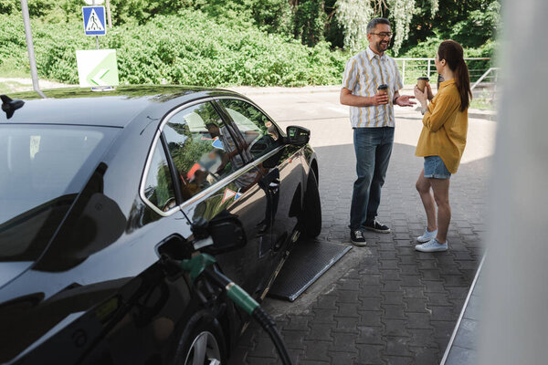 Selective focus of smiling man holding paper cup near wife while fueling car on gas station 