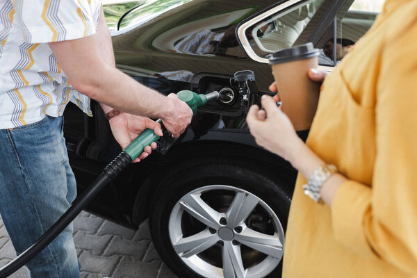 Cropped view of man holding fueling nozzle near gas tank of car near woman with paper cup on gas station 