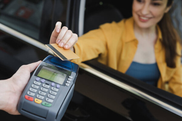 Selective focus of smiling woman holding credit card while sitting in car near worker of gas station holding payment terminal