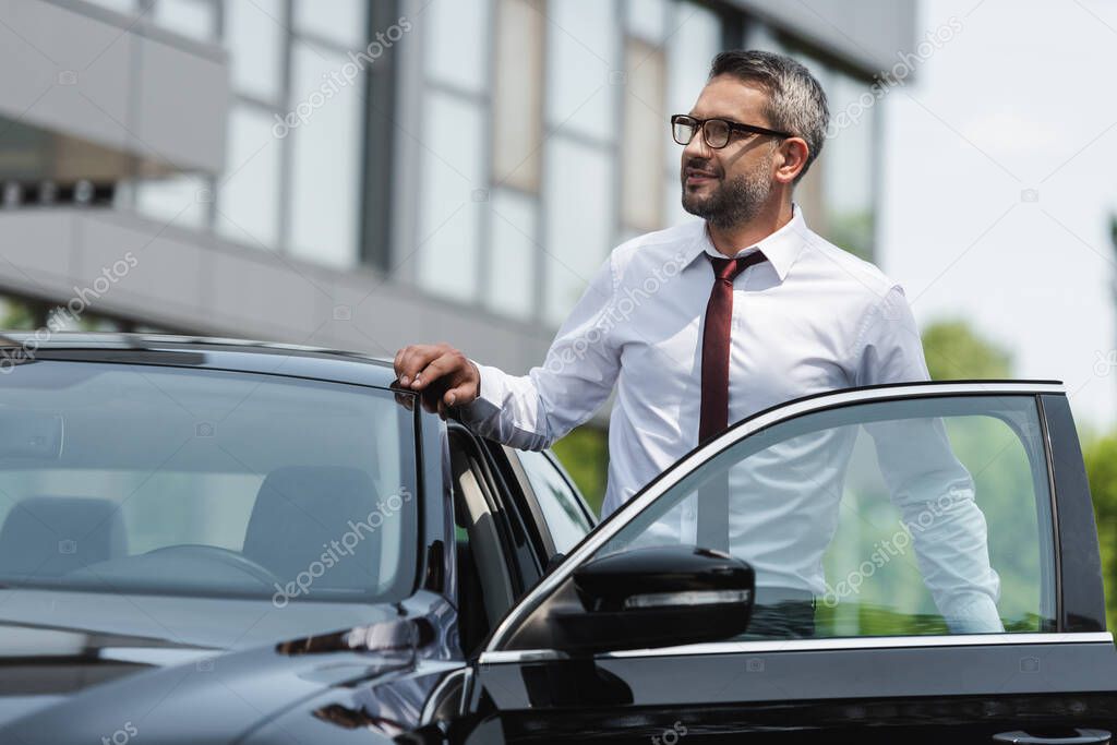 Selective focus of handsome businessman standing near car with open door on urban street 