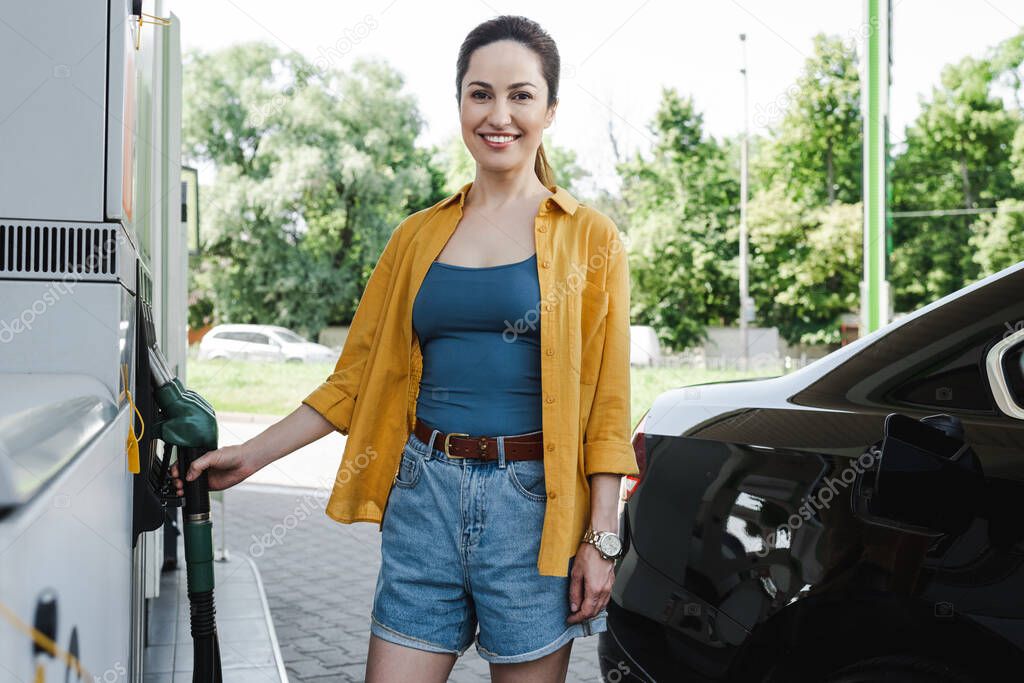 Selective focus of beautiful smiling woman holding fueling nozzle near car on gas station 