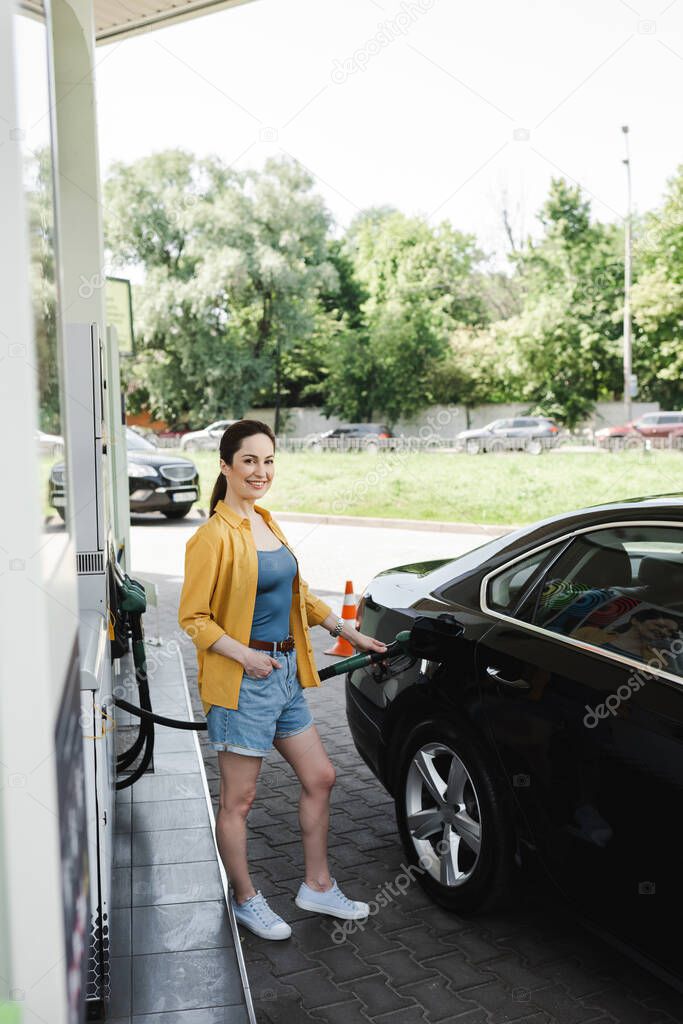 Selective focus of smiling woman with hand in pocket looking at camera and fueling car on gas station 