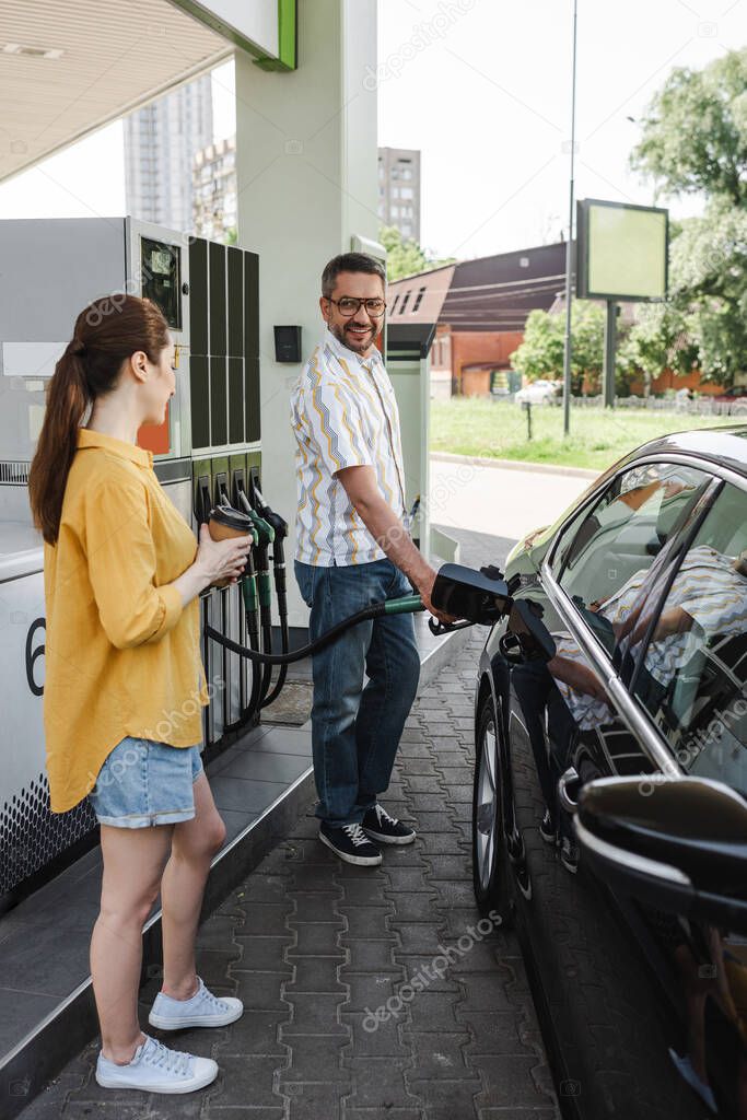Selective focus of smiling man fueling car near wife with coffee to go on gas station 