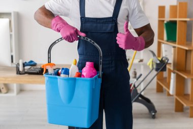 Cropped view of African american cleaner showing thumb up and holding bucket of detergents in office  clipart