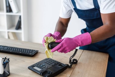 Ofis masasında telefon temizlerken bez kullanan Afro-Amerikan temizlikçi görüntüsü. 