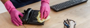 Panoramic crop of african american cleaner in rubber gloves cleaning telephone wit rag in office  clipart