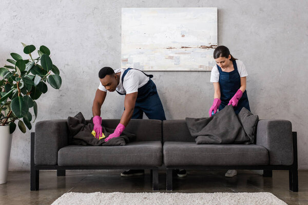 Multicultural cleaners in uniform cleaning couch with rags in living room