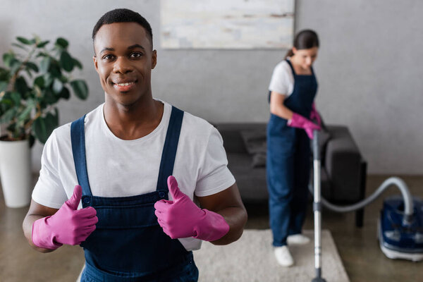 Selective focus of smiling african american cleaner showing thumbs up near colleague cleaning carpet 