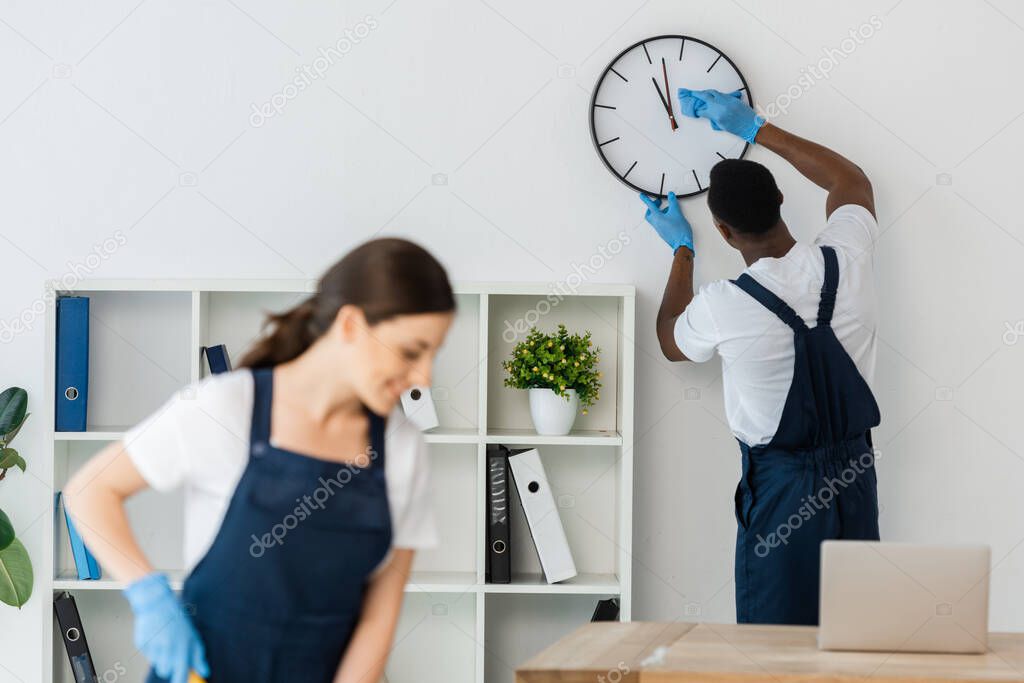 Selective focus of african american cleaner cleaning clock near smiling colleague in office