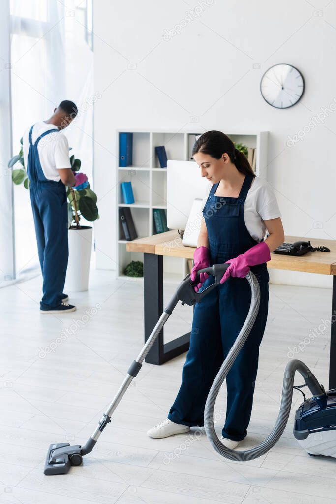 Selective focus of cleaner using vacuum cleaner near african american colleague in office 