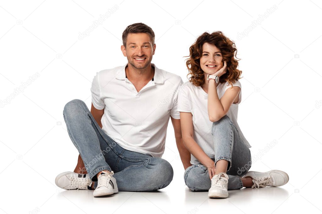 happy couple in denim jeans sitting and looking at camera on white