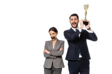 excited businessman holding trophy near businesswoman with duct tape on mouth standing with crossed arms isolated on white clipart