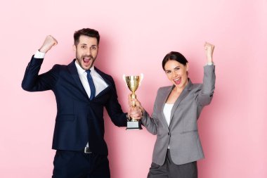 excited businessman and businesswoman holding trophy and gesturing on pink, gender equality concept clipart