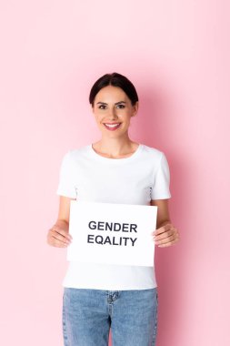 happy woman in white t-shirt holding placard with gender equality lettering on pink  clipart
