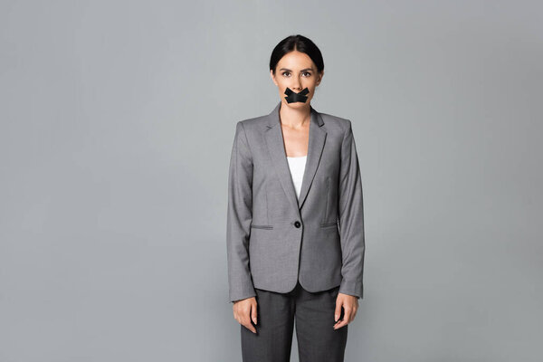 businesswoman with scotch tape on mouth standing isolated on grey