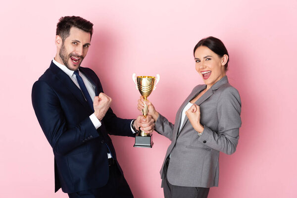 excited businessman and businesswoman in suits holding trophy and gesturing on pink, gender equality concept