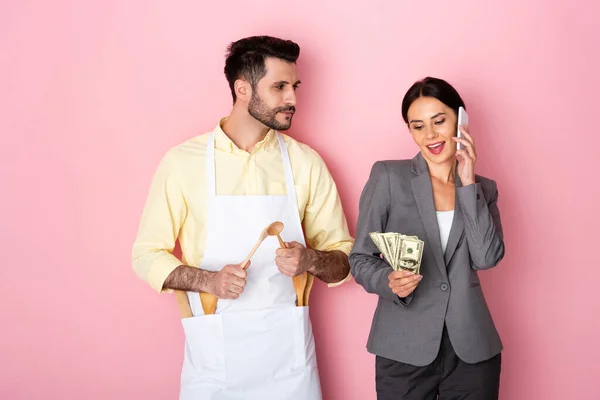 エプロンのハンサムな男が木製のスプーンを保持し ピンクの上にスマートフォンで話してお金でビジネス女性を見て — ストック写真