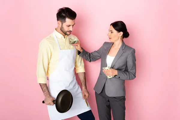 businesswoman putting money in pocket of handsome man in apron holding frying pan on pink