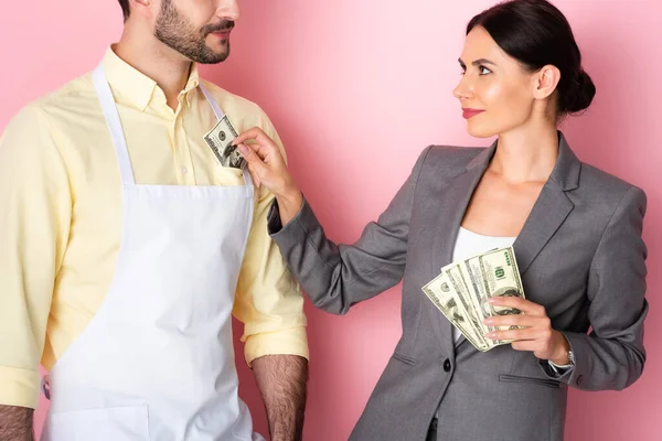 businesswoman putting money in pocket of bearded man in apron on pink