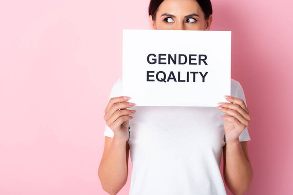 woman in white t-shirt covering face with gender equality lettering on placard and looking away on pink 