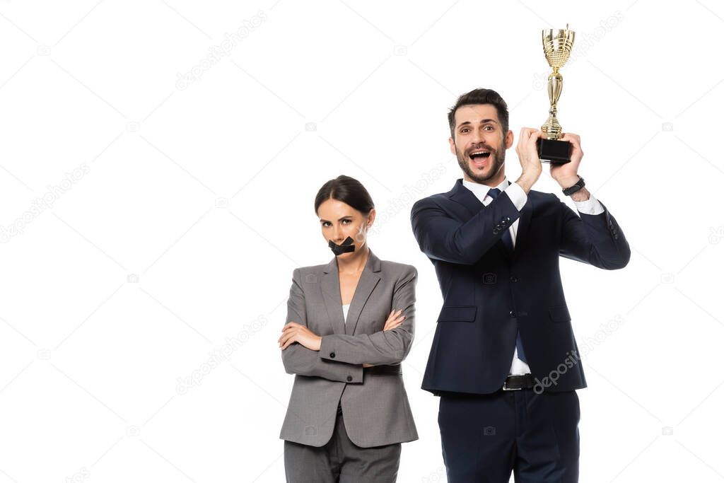 excited businessman holding trophy near businesswoman with duct tape on mouth standing with crossed arms isolated on white