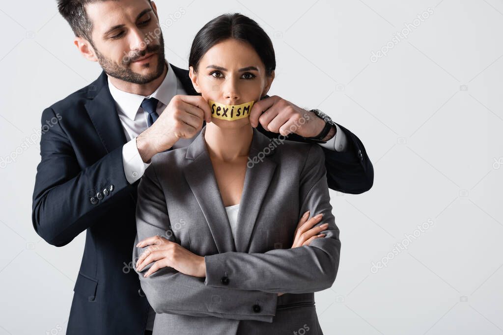 businessman touching scotch tape with sexism lettering on mouth of businesswoman standing with crossed arms isolated on white