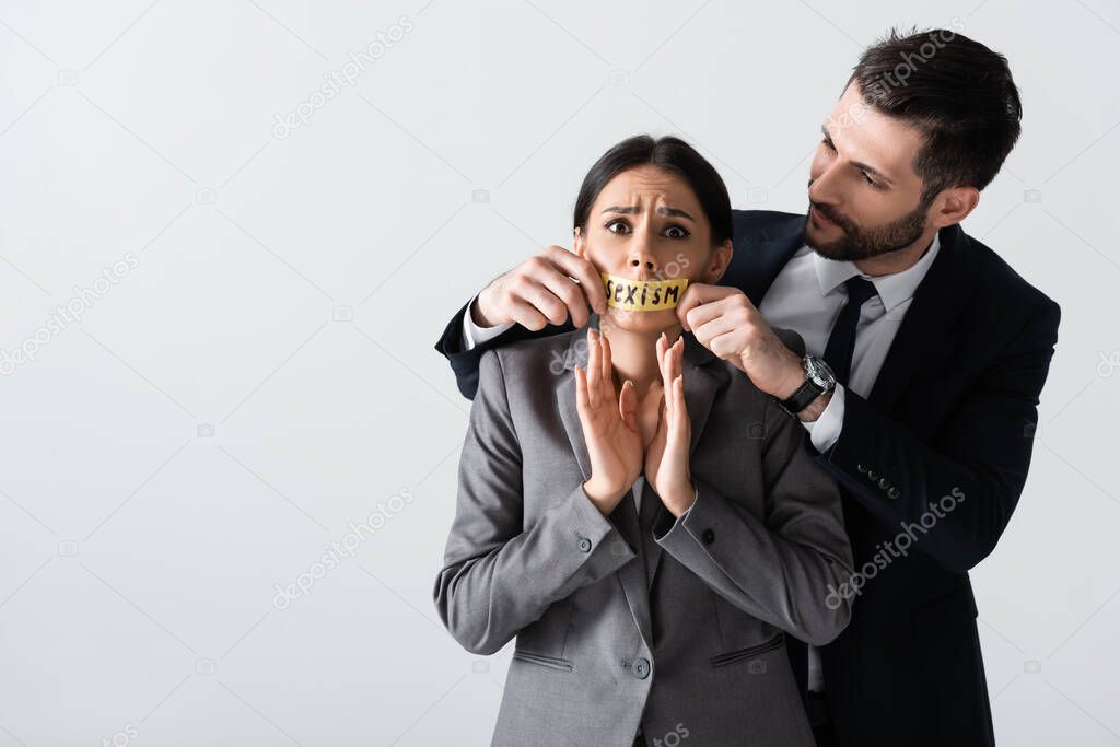 businessman touching scotch tape with sexism lettering on mouth of scared businesswoman gesturing isolated on white