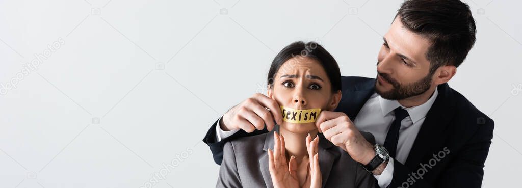 horizontal image of businessman touching scotch tape with sexism lettering on mouth of scared businesswoman gesturing isolated on white