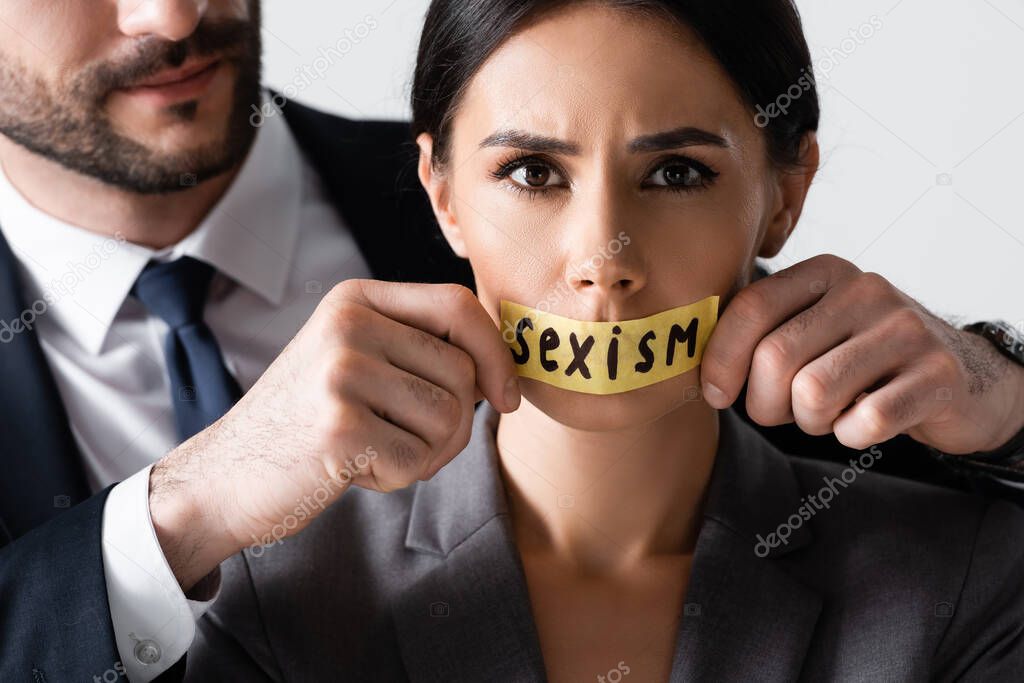 bearded businessman touching scotch tape with sexism lettering on mouth of businesswoman looking at camera isolated on white