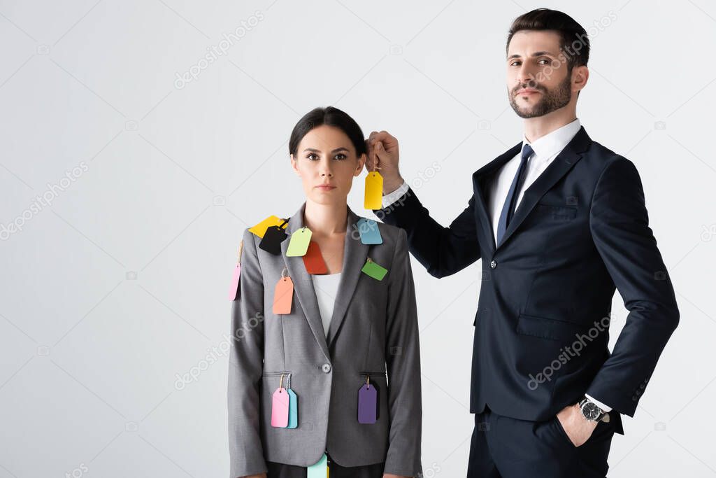 bearded businessman standing with hand in pocket and putting label on beautiful businesswoman isolated on white, sexism concept 