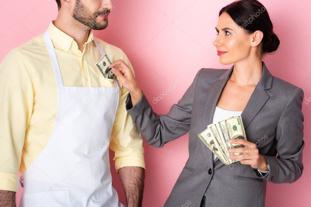 businesswoman putting money in pocket of bearded man in apron on pink 