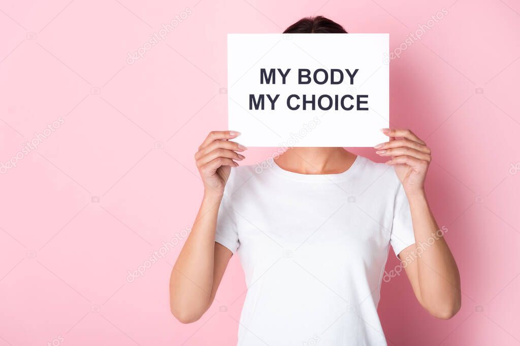 woman in white t-shirt holding placard with my body my choice lettering while covering face on pink 