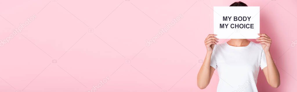 panoramic orientation of woman in white t-shirt holding placard with my body my choice lettering while covering face on pink 