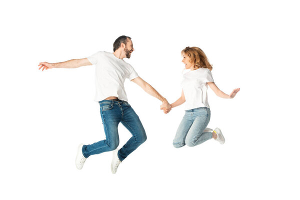 happy adult couple in white t-shirts holding hands while jumping isolated on white