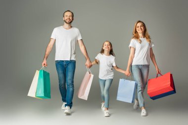 Neşeli aile renkli alışveriş torbalarıyla yürüyor ve gri arka planda el ele tutuşuyorlar.
