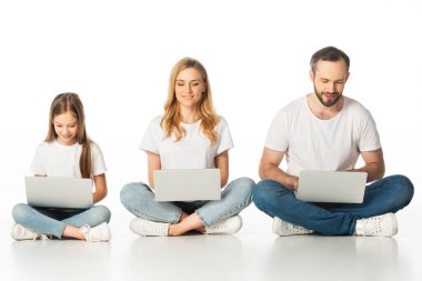 Neşeli bir aile dizüstü bilgisayarlarıyla yerde oturuyor çapraz bacaklarıyla beyazlar içinde.