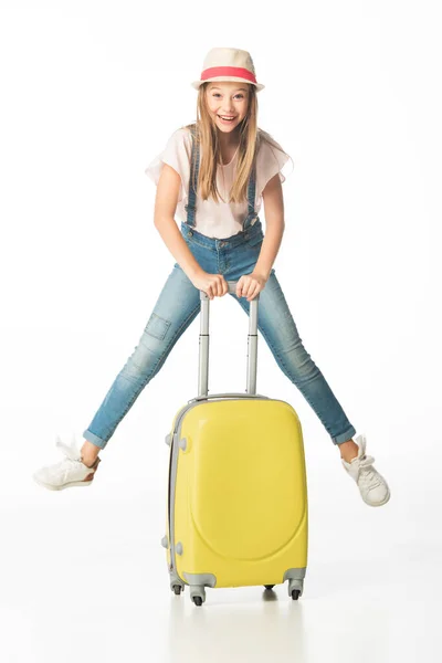戴帽子快乐的女孩跳到黄色旅行袋边 与白色隔离 — 图库照片