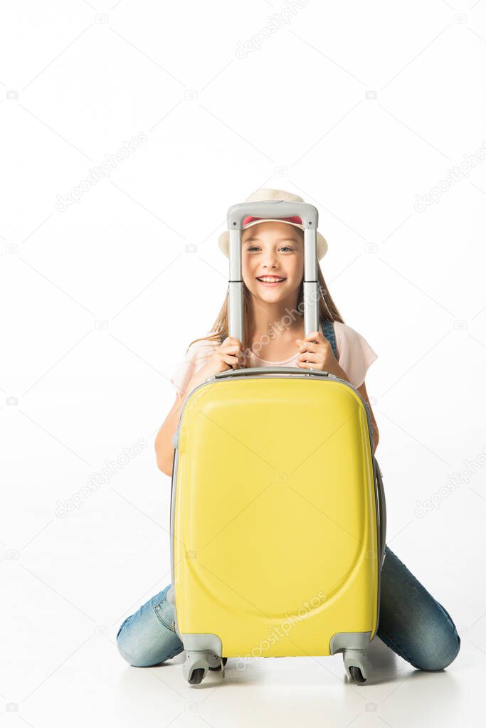 cheerful girl in hat near yellow travel bag isolated on white