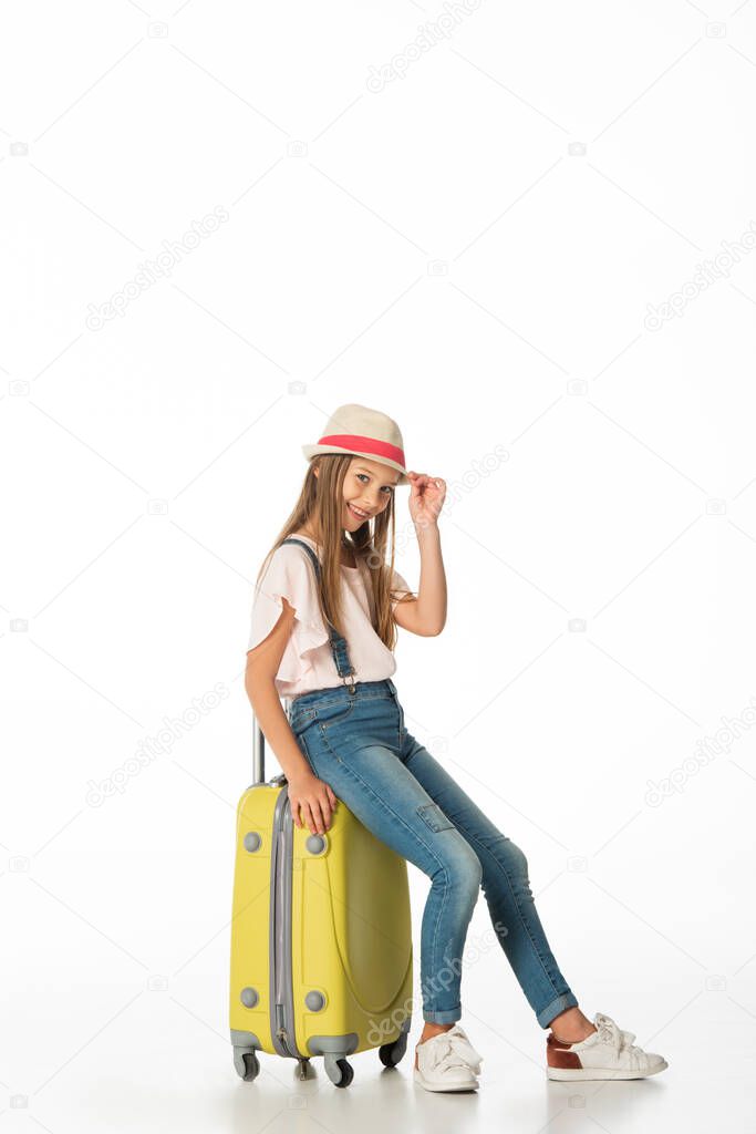 cheerful girl in hat on yellow travel bag isolated on white