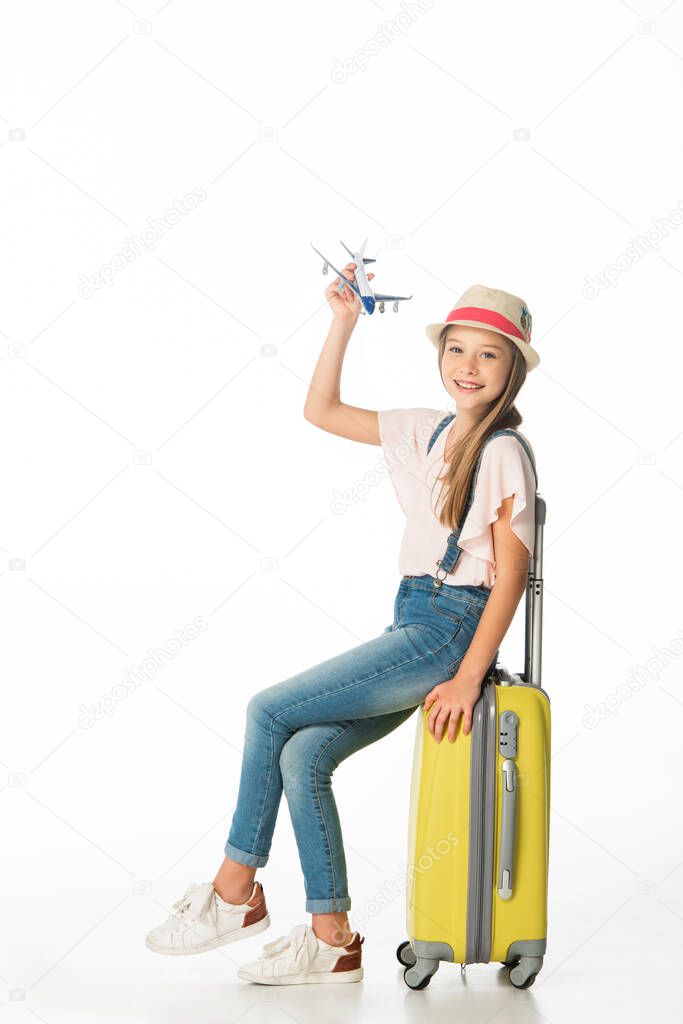 cheerful girl in hat with plane model on yellow travel bag isolated on white