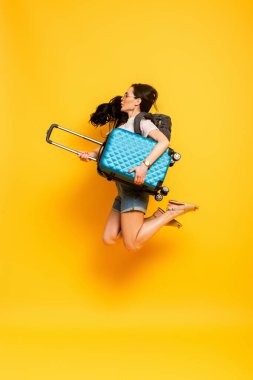 side view of brunette woman jumping with backpack and suitcase on yellow background clipart