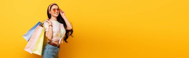 smiling stylish summer brunette girl with shopping bags on yellow background, panoramic shot clipart