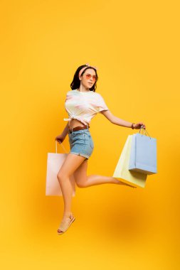 full length view of stylish summer brunette girl jumping with shopping bags on yellow background clipart