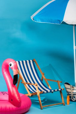 striped deck chair near inflatable flamingo, sunscreen, beach bag and umbrella on blue background clipart