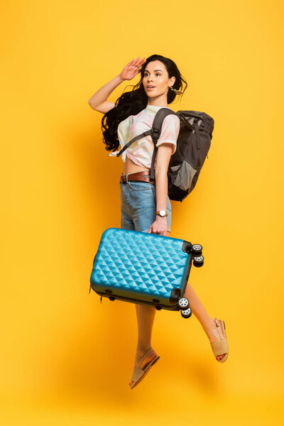 brunette woman jumping with backpack and suitcase and looking away on yellow background