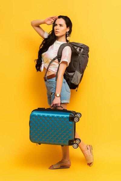 brunette woman with backpack and suitcase looking away on yellow background