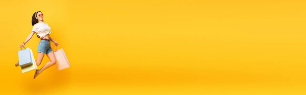 full length view of stylish summer brunette girl jumping with shopping bags on yellow background, panoramic shot