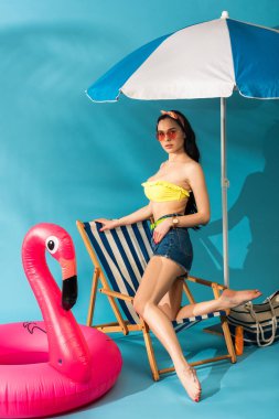 Havalı kız güvertede poz veriyor şişirilebilir flamingo, kumsal çantası ve mavi arka planda şemsiye.