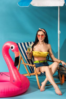 Gülümseyen stil sahibi kız güverte koltuğunda şemsiyenin altında mavi arka planda şişme flamingo altında kokteyl ile oturuyor.